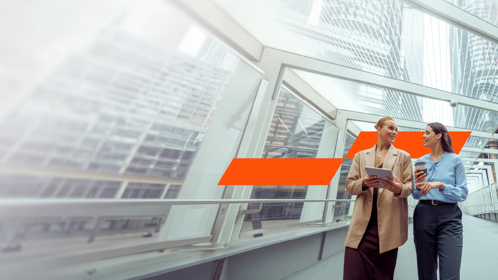 Two professional women walk and converse along a modern glass-walled pedestrian bridge with a cityscape.