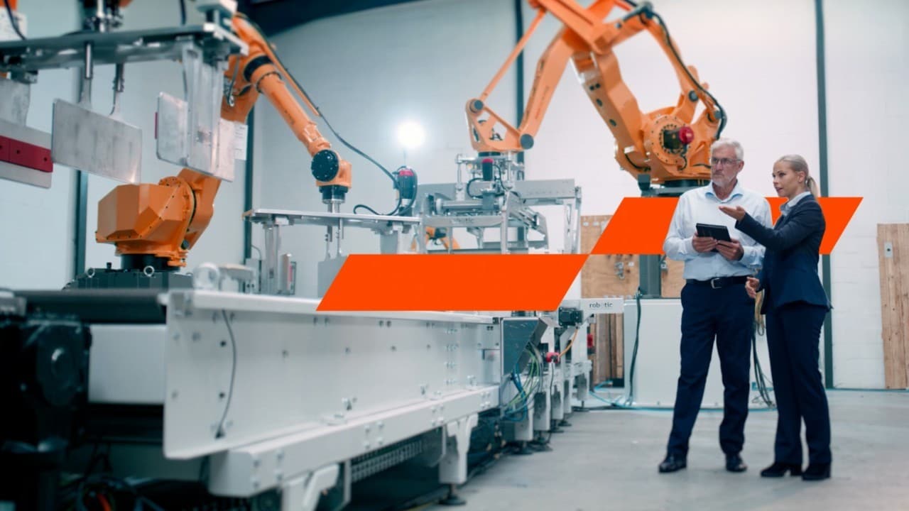 Two professionals discussing and reviewing a tablet near robotic arms in a modern industrial automation facility.
