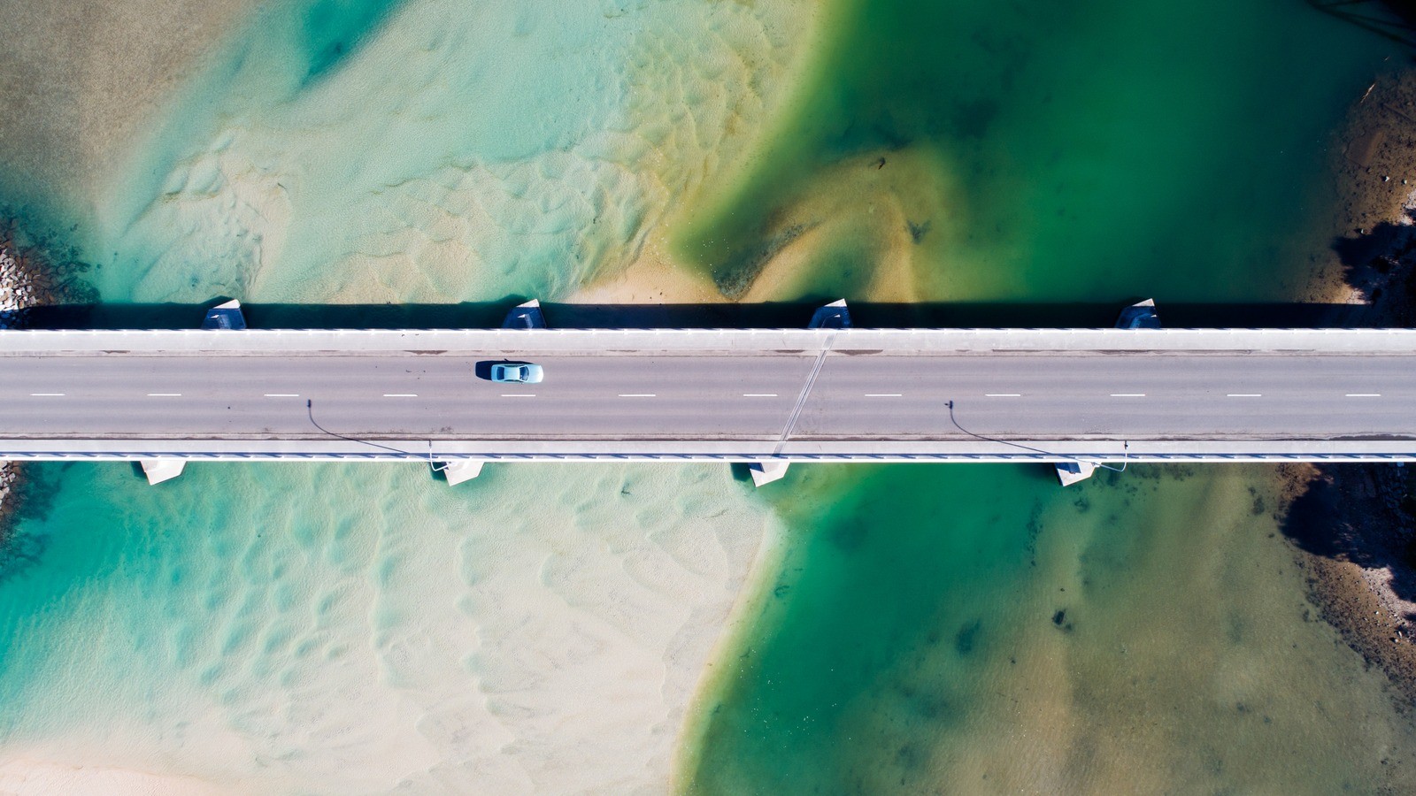 Bridge stretches over shallow turquoise water as lone blue car crosses