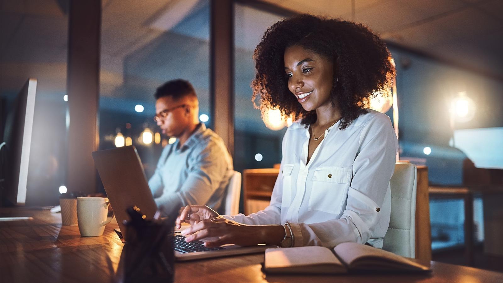 Young businesswoman working on a laptop alongside her colleague in an office at night
